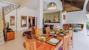 A spacious dining area with a wooden table set for four, surrounded by wooden chairs. A staircase is visible to the left, leading to an upper level. - Villa Bali Caviar (Seminyak)