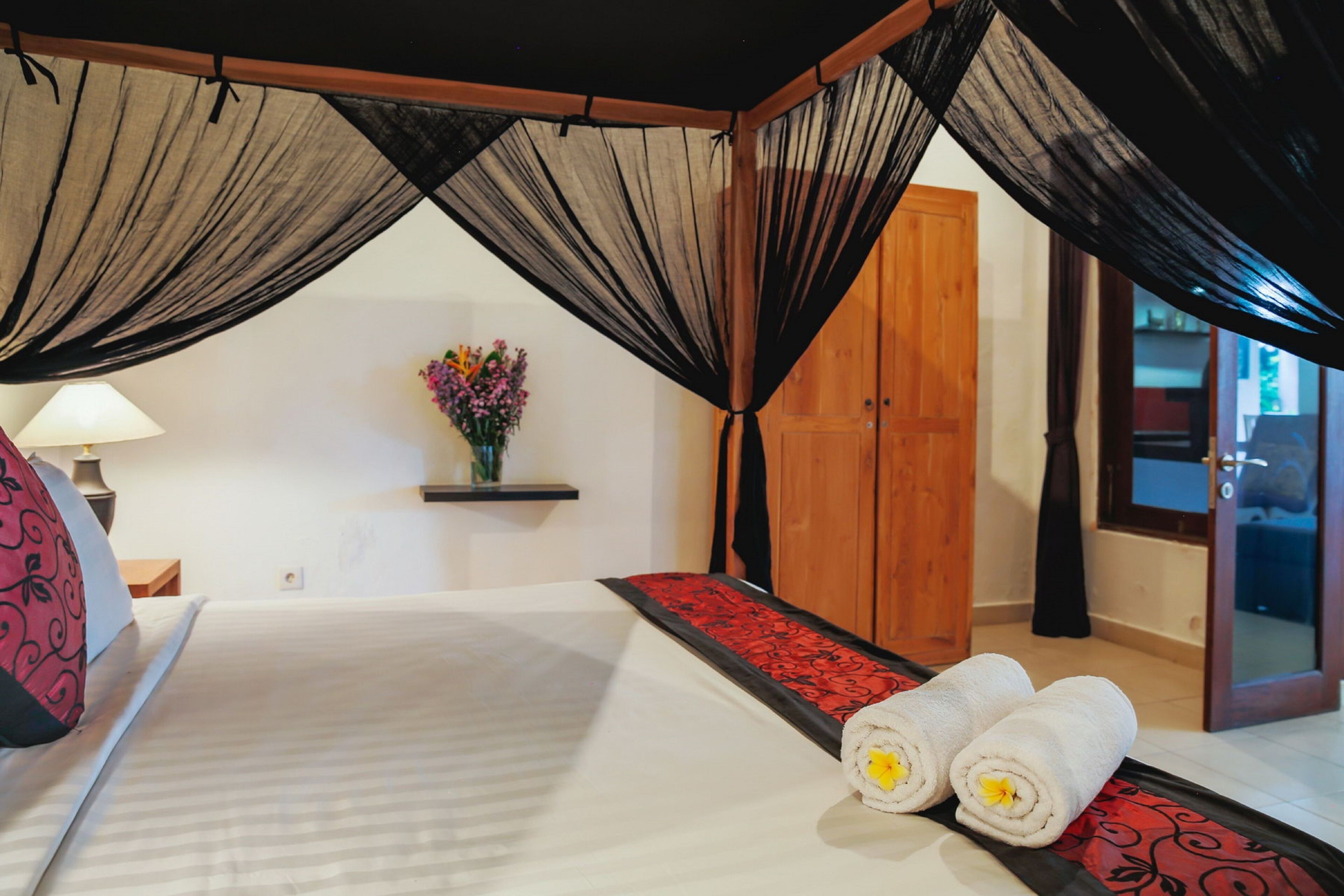 A hotel room with a bed, a nightstand, a lamp, a vase with flowers, and a wooden door.