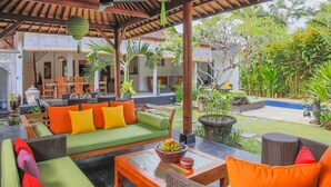 A covered outdoor seating area with a green sofa, orange cushions, and a table with a bowl of fruit. - Villa Bali Caviar (Seminyak)