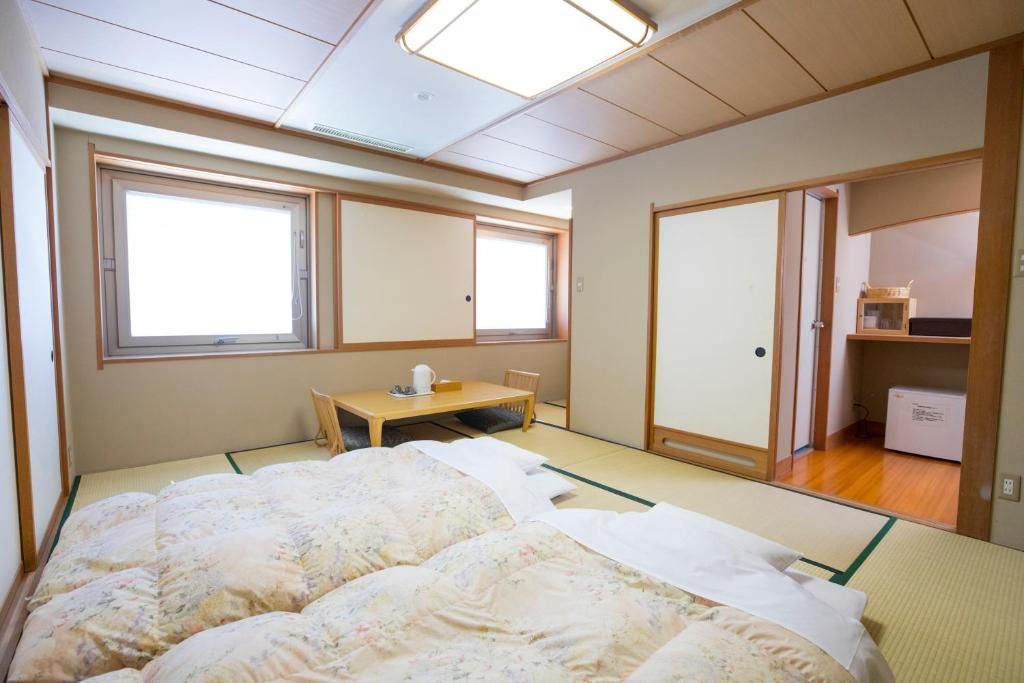 Une pièce de style japonais traditionnel, avec un sol recouvert de tatamis, un lit avec une literie à motifs floraux, une petite table avec une bouilloire et des portes coulissantes donnant sur une autre pièce.