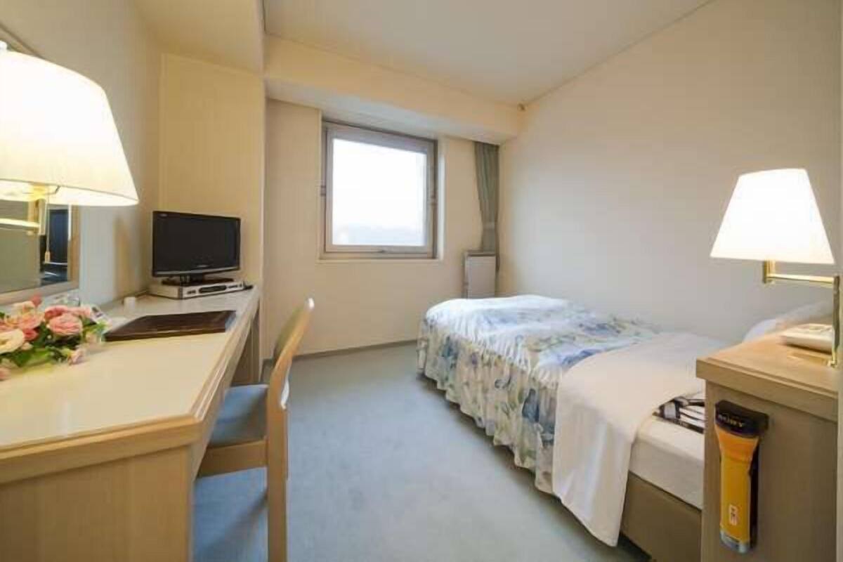 A hotel room with a bed, a desk with a computer and lamp, a window, and a small table with a lamp.
