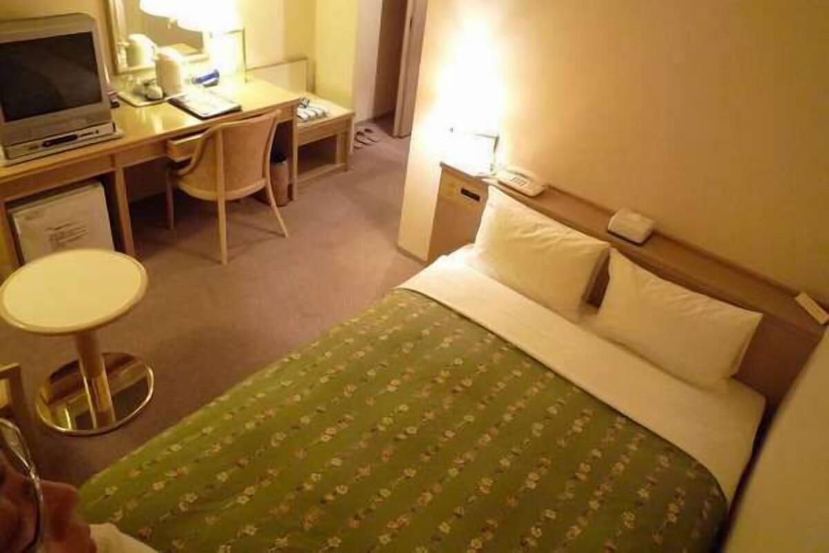 A hotel room with a bed, a desk with a computer, a chair, a small table, and a mirror.