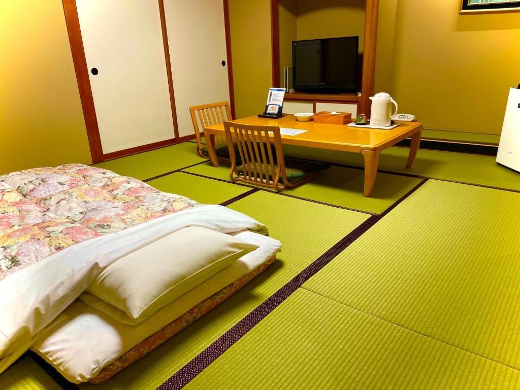 A room with tatami flooring, a bed with floral bedding, a wooden table with a laptop, a television, and a small refrigerator.