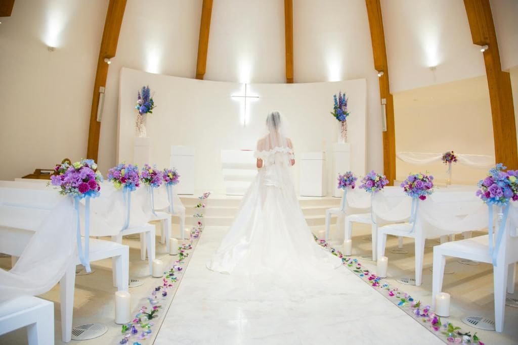 A church interior with white chairs arranged in rows, a central aisle, and floral decorations.