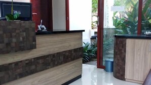 A reception area with a wooden reception desk, a computer monitor, and a glass partition with plants. - D'Pande (Kuta)
