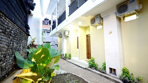 A narrow courtyard with a stone wall, potted plants, and a pathway leading to a building with balconies and air conditioning units. - D'Pande (Kuta)