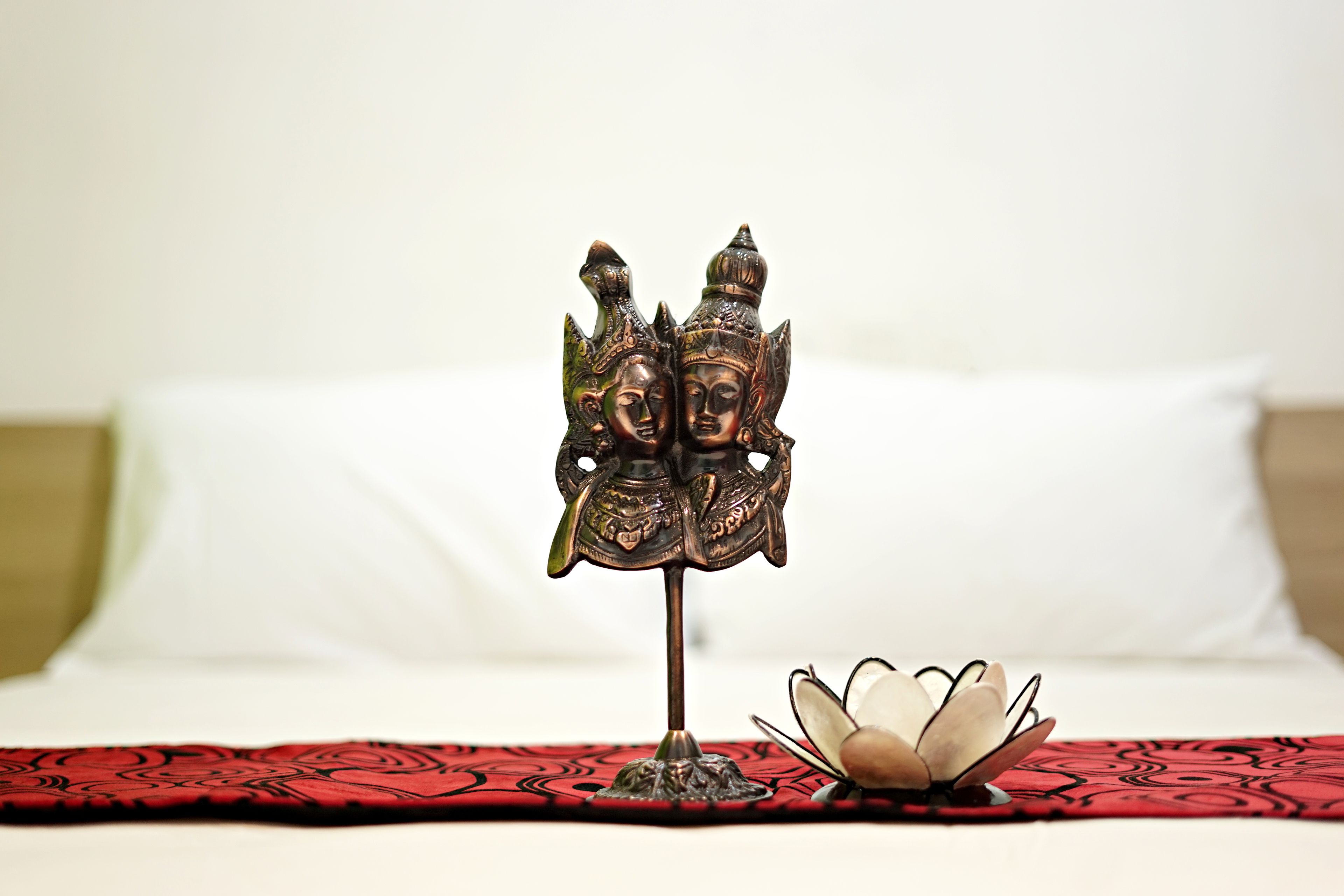 A bed with a red and black patterned bedspread, a decorative metal object with faces, and a lotus flower on a small stand.