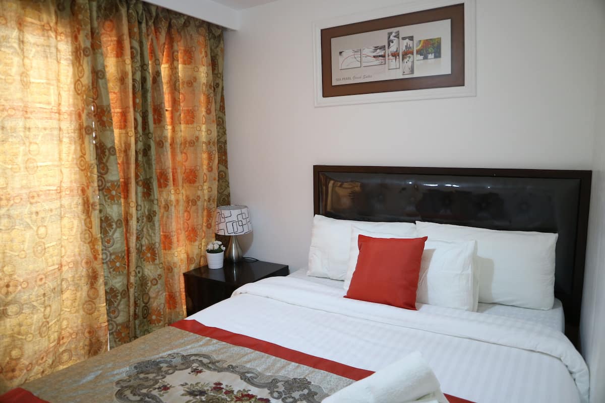 A neatly made bed with white linens and a red accent pillow, a nightstand with a lamp, and a framed artwork on the wall.