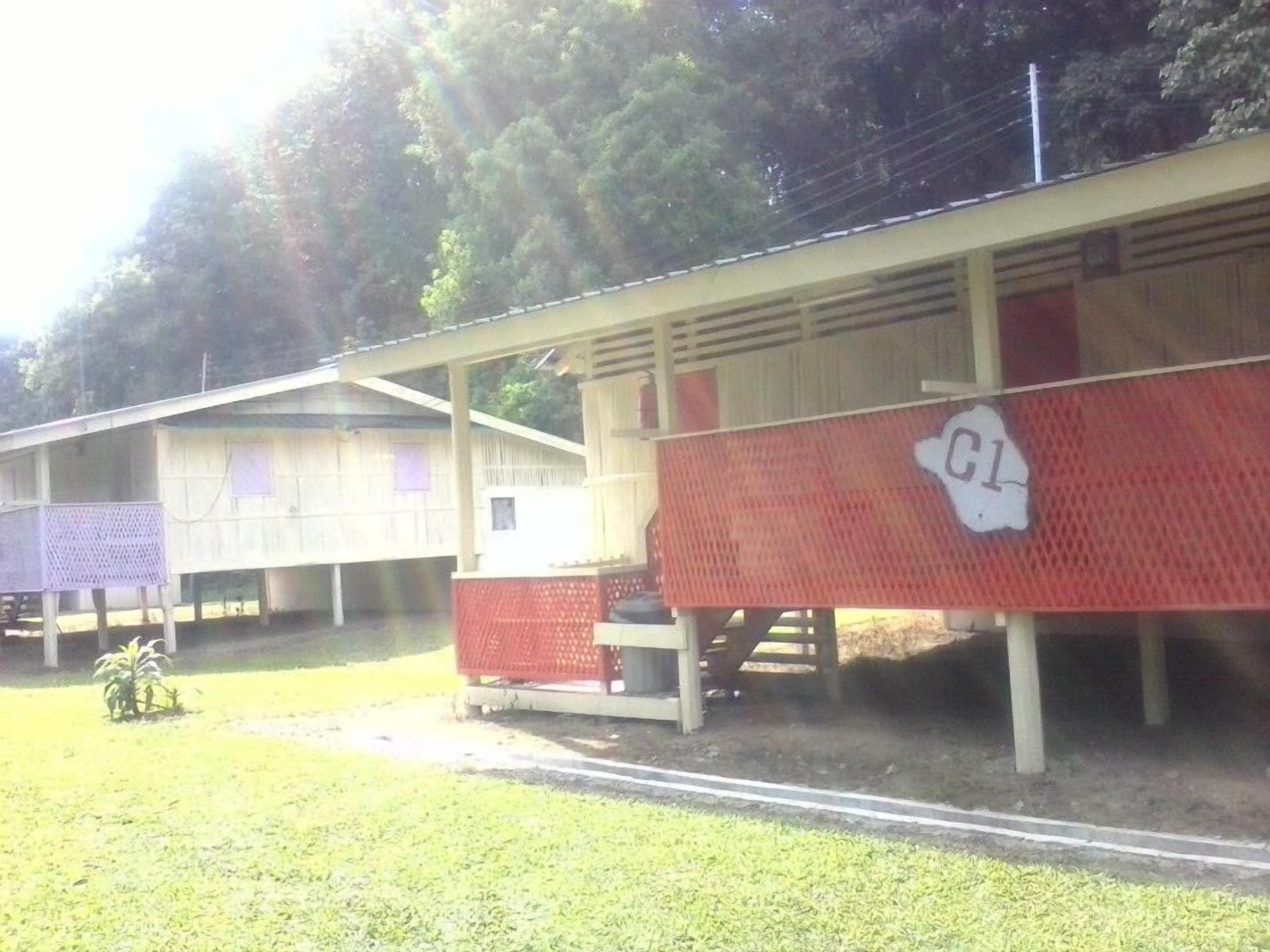Een rij eenvoudige, gelijkvloerse houten hutten in een rood-wit kleurenschema, elk met een klein terras en een bordje met 'C1' erop.