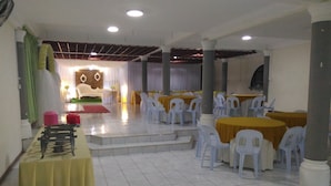 Un salón de banquetes con suelo de baldosas blancas, mesas redondas con manteles amarillos y sillas de plástico blanco.