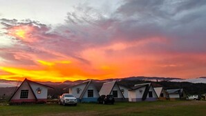 Una hilera de cabañas sencillas de un solo nivel con techos a dos aguas, frente a un paisaje de montañas y un cielo al atardecer.