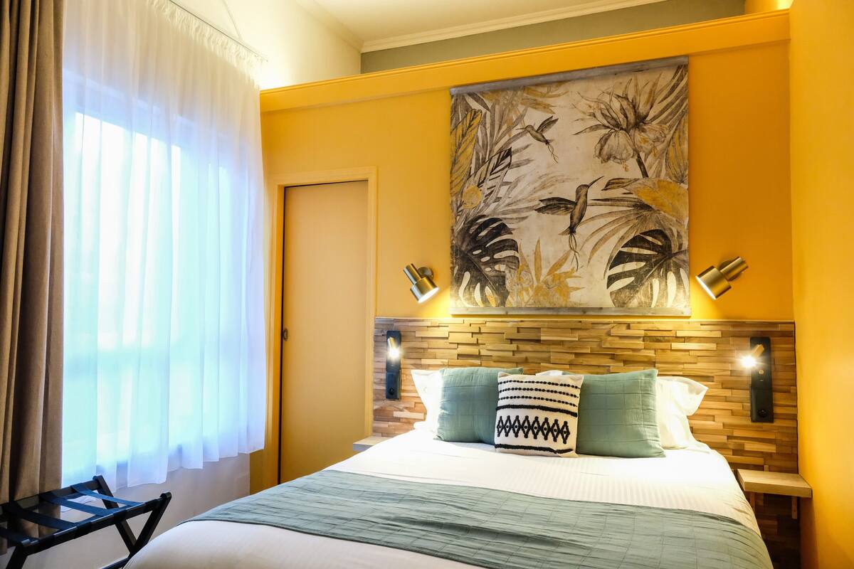 A hotel room with a bed, a headboard with a wooden panel design, a wall-mounted artwork depicting tropical leaves and birds, and a nightstand with a lamp.