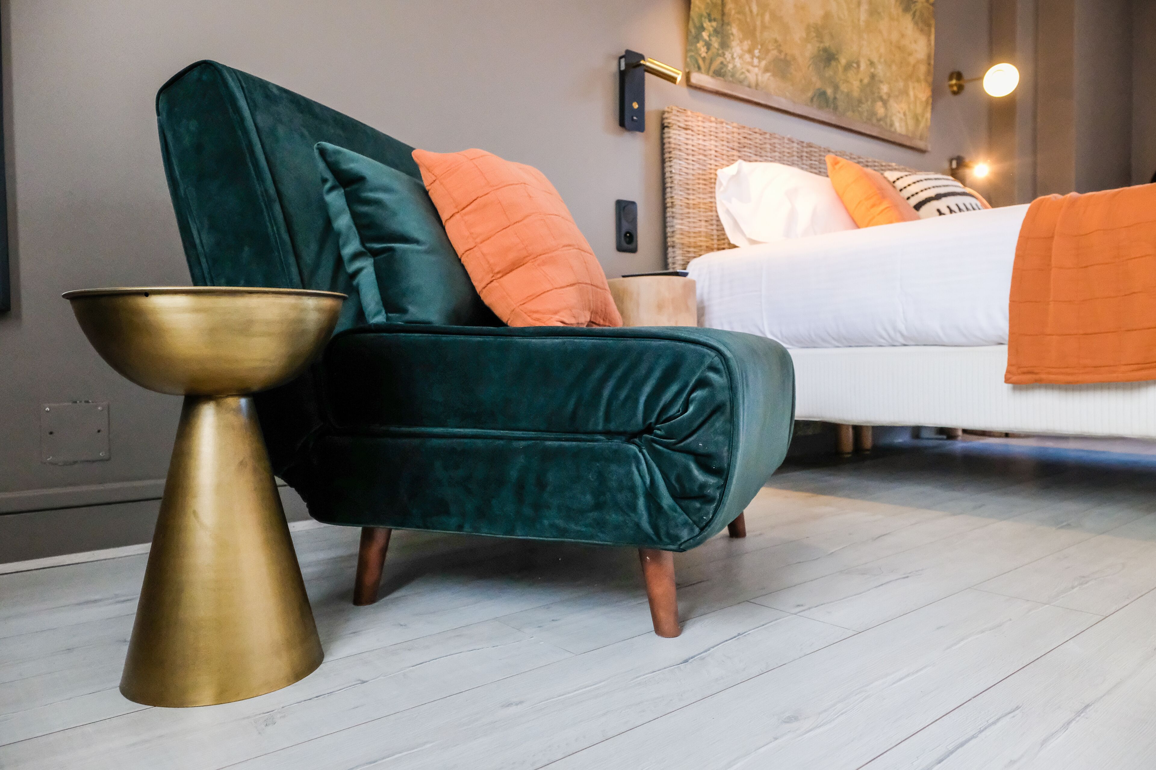 A modern bedroom with a green velvet armchair, a brass side table, and a white bed with orange and white pillows.