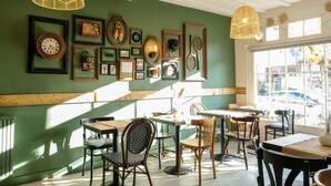 Ein gemütliches Café mit Holzmöbeln, gerahmten Bildern an der Wand und großen Fenstern, die viel Tageslicht hereinlassen.