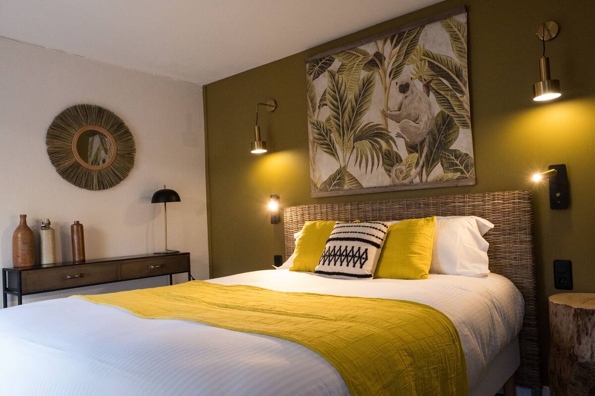 A neatly made bed with a yellow blanket, a headboard with two pillows, a wall-mounted mirror, and two wall sconces.