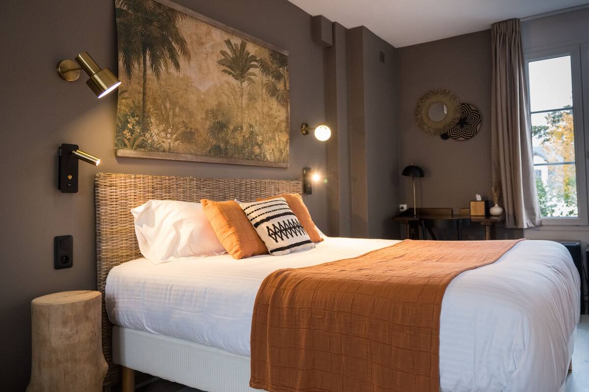 A neatly made bed with a white and brown bedspread, a woven headboard, and two pillows. There is a wall-mounted light fixture and a painting of tropical foliage above the bed.