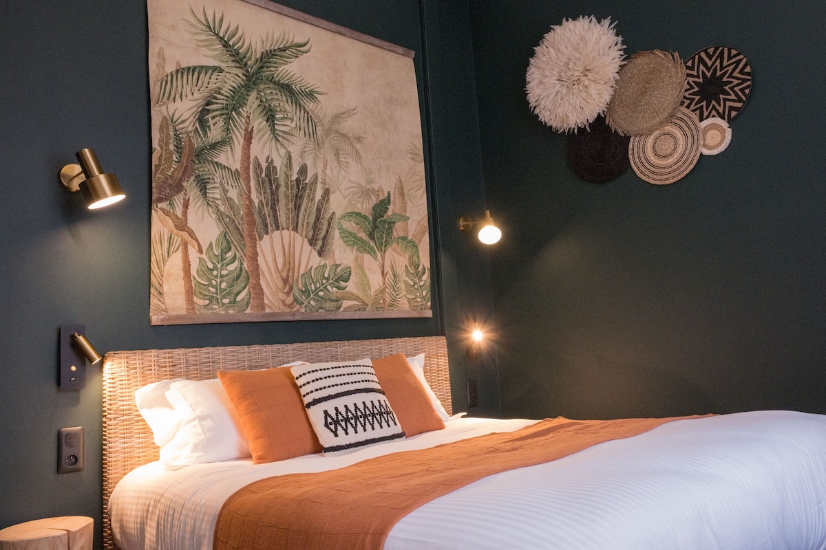 A neatly made bed with a white and brown blanket, a tropical-themed painting on the wall, and a wall-mounted light fixture.