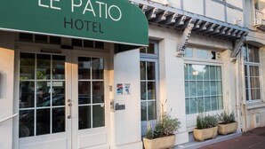 Das Außere des Le Patio Hotels mit einer grünen Markise, großen Fenstern und Pflanzkübeln am Eingang.