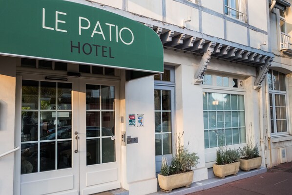 Das Außere des Le Patio Hotels mit einer grünen Markise, großen Fenstern und Pflanzkübeln am Eingang.