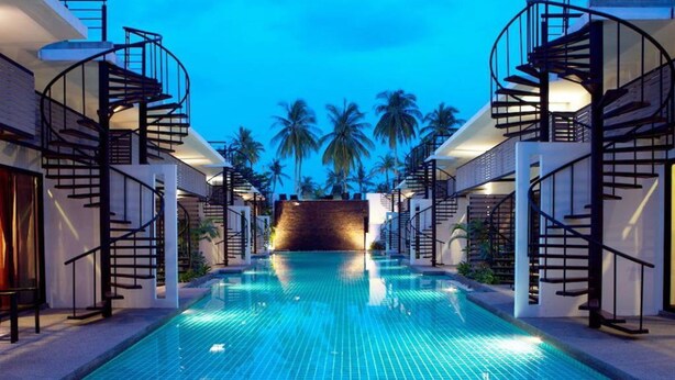 A swimming pool with a modern design, surrounded by buildings with spiral staircases and palm trees in the background. - The Kris Resort Condotel at Bagtao Beach (Choeng Thale)