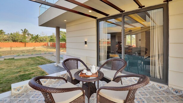 A balcony with wicker furniture and a table set with tea and cups, overlooking a grassy area and a building with large glass doors. - Goverdhan Greens Resort Dwarka (Dwarka)