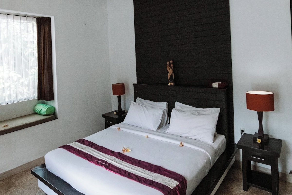 A bedroom with a large bed, two bedside tables, a window with curtains, and a dark headboard.