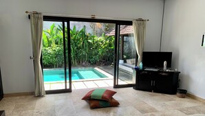 Una habitación con una puerta corrediza de vidrio que da a una piscina, un televisor de pantalla plana sobre un mueble negro y una pequeña zona de estar con cojines.