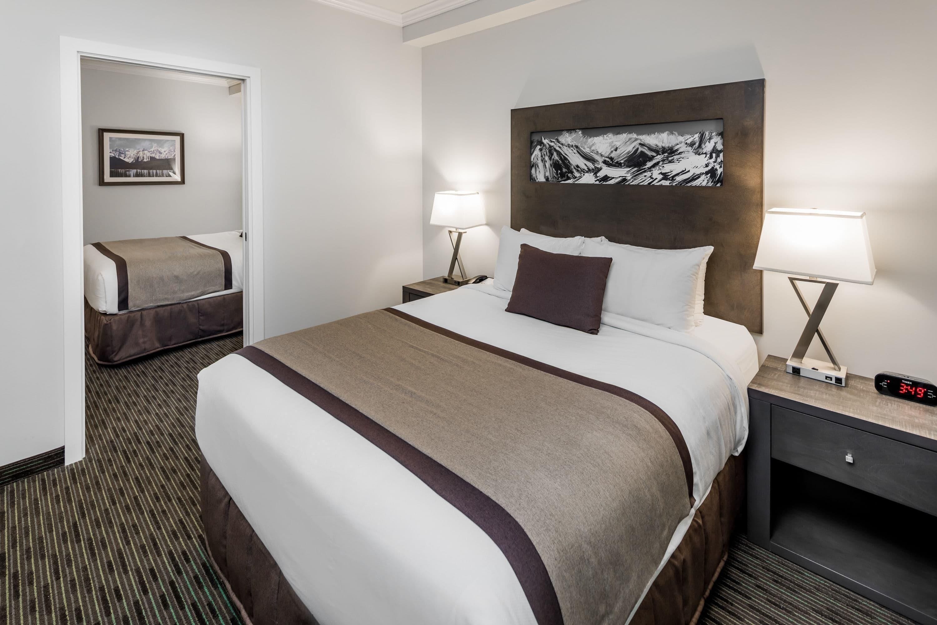 Habitación de hotel con una cama grande, dos mesitas de noche con lámparas, una mesita auxiliar y un cuadro enmarcado en la pared, además de una puerta que da a otra habitación.