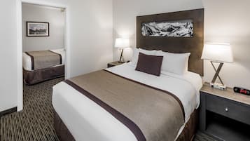 Habitación de hotel con una cama grande, dos mesitas de noche con lámparas, una mesita auxiliar y un cuadro enmarcado en la pared, además de una puerta que da a otra habitación.