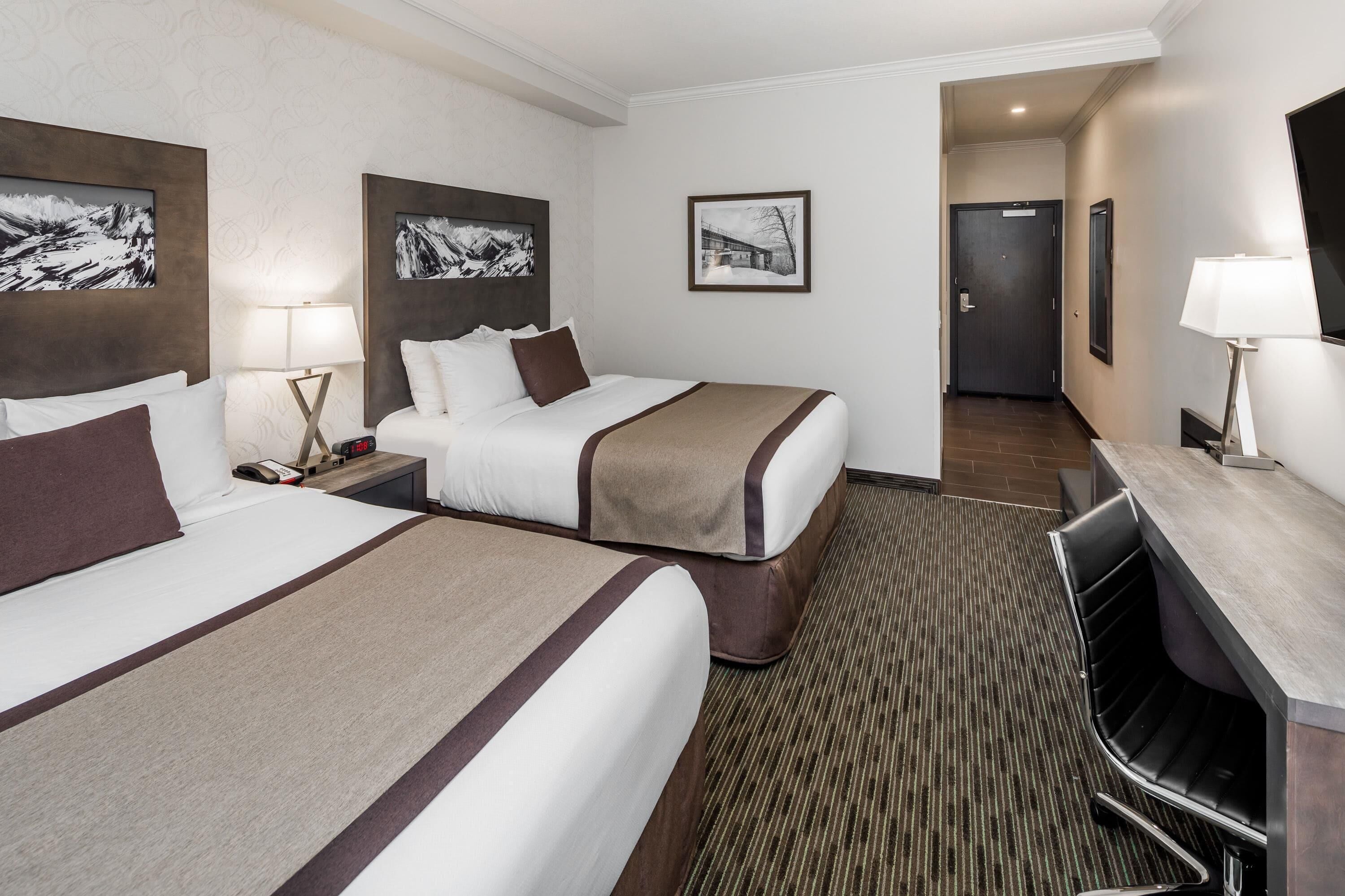 A hotel room with two beds, a desk, a chair, a TV, and framed pictures on the walls.