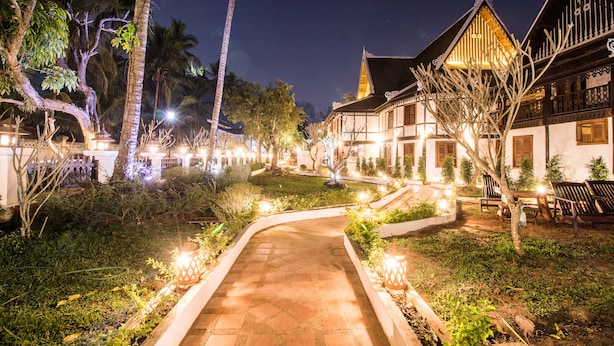 Un sendero bien iluminado conduce a una edificación tradicional con techo de tejas y balcones de madera, rodeada de exuberante vegetación y palmeras.