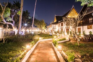 A well-lit pathway leading to a traditional building with a tiled roof and wooden balconies, surrounded by lush greenery and palm trees. - Royal Peacock Boutique Hotel (Luang Prabang)