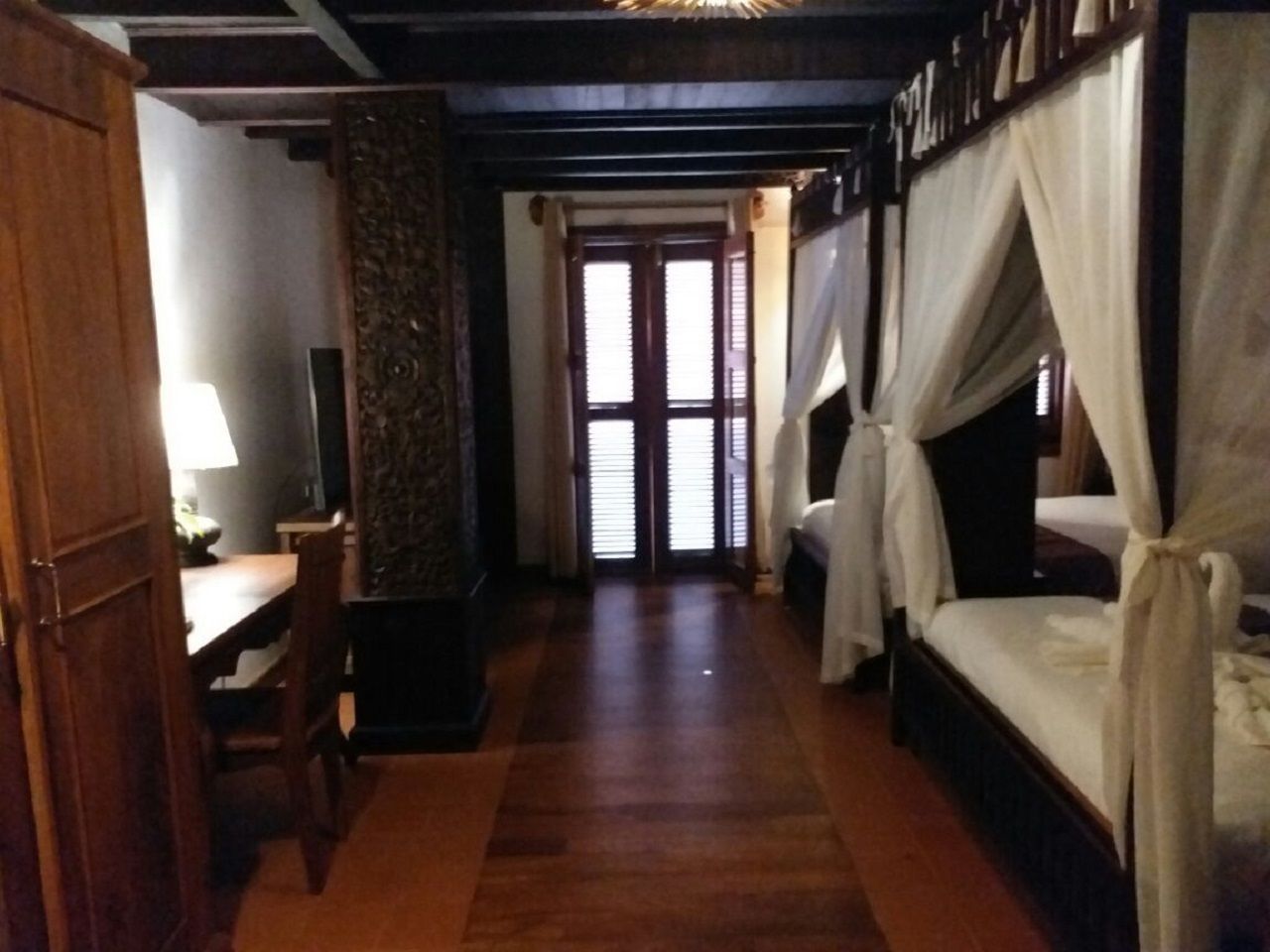Una habitación con piso de madera, un escritorio con lámpara, una cama con dosel y una puerta que da a otra habitación.