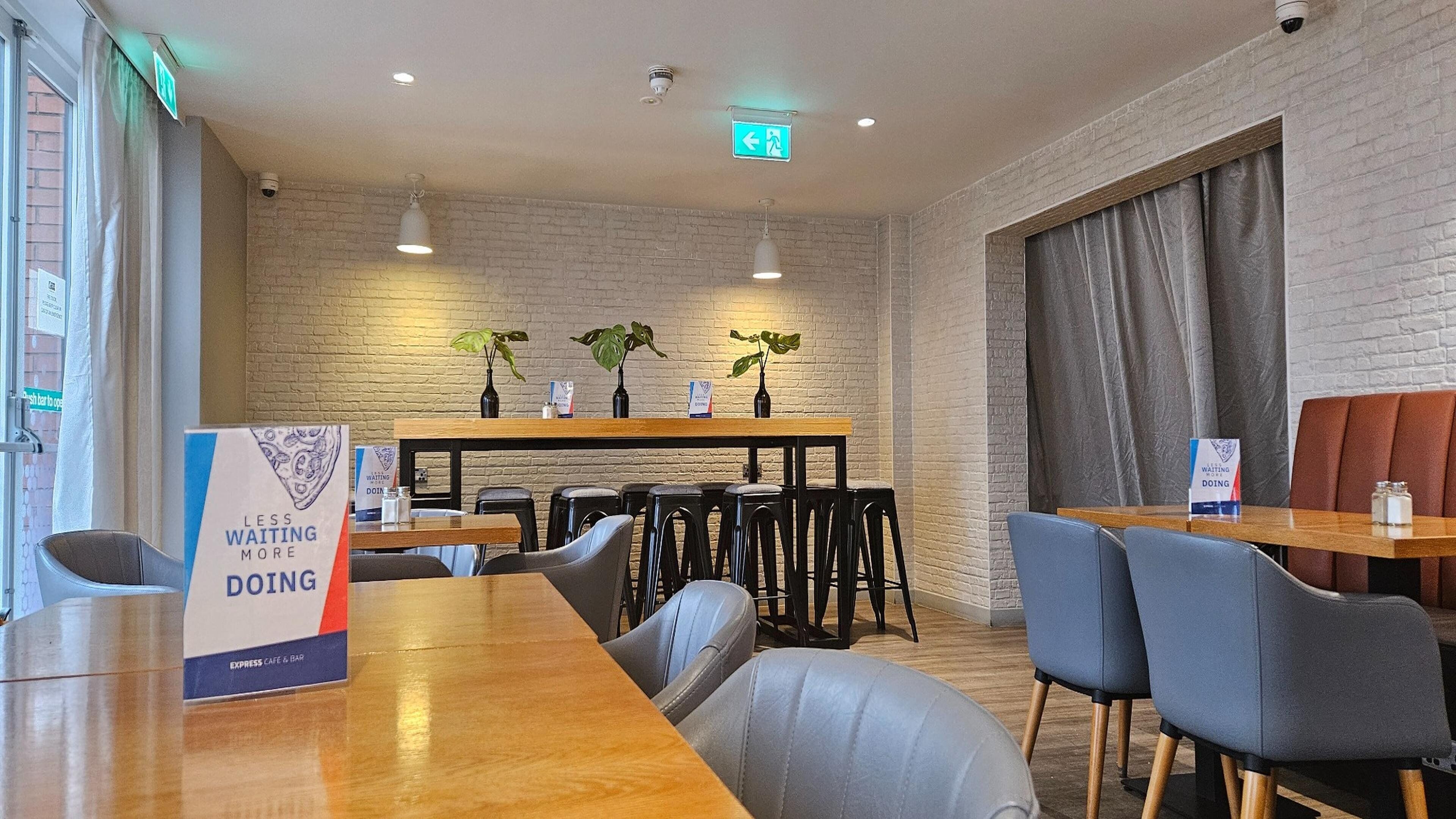 A modern cafe with wooden tables and chairs, a brick wall, and a sign indicating a waiting area.