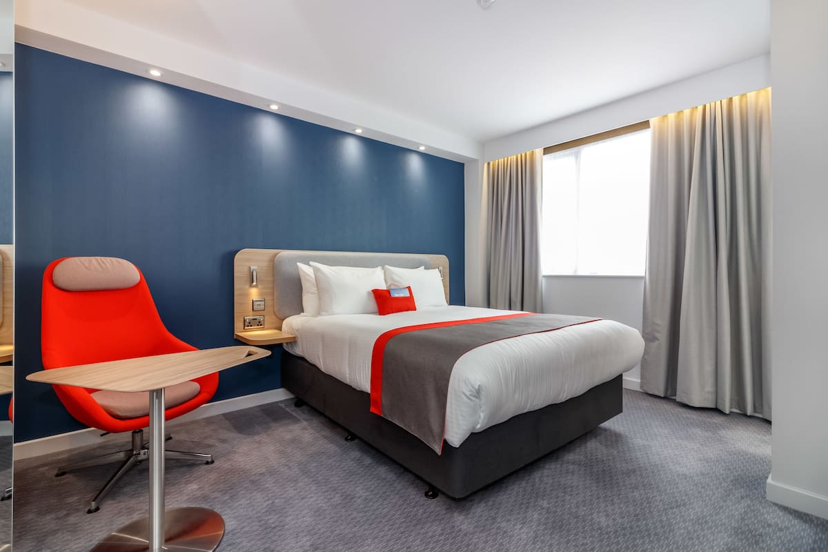 A modern hotel room with a bed, a desk, a chair, and a large window with curtains.