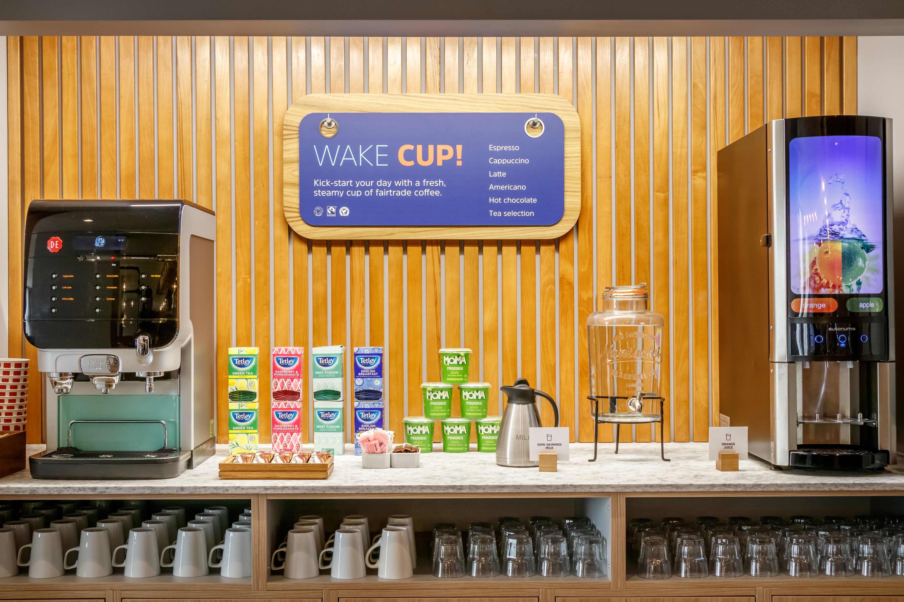 A coffee station with a sign reading "WAKE CUP!" offering various coffee options like espresso, cappuccino, latte, Americano, hot chocolate, and tea selection.