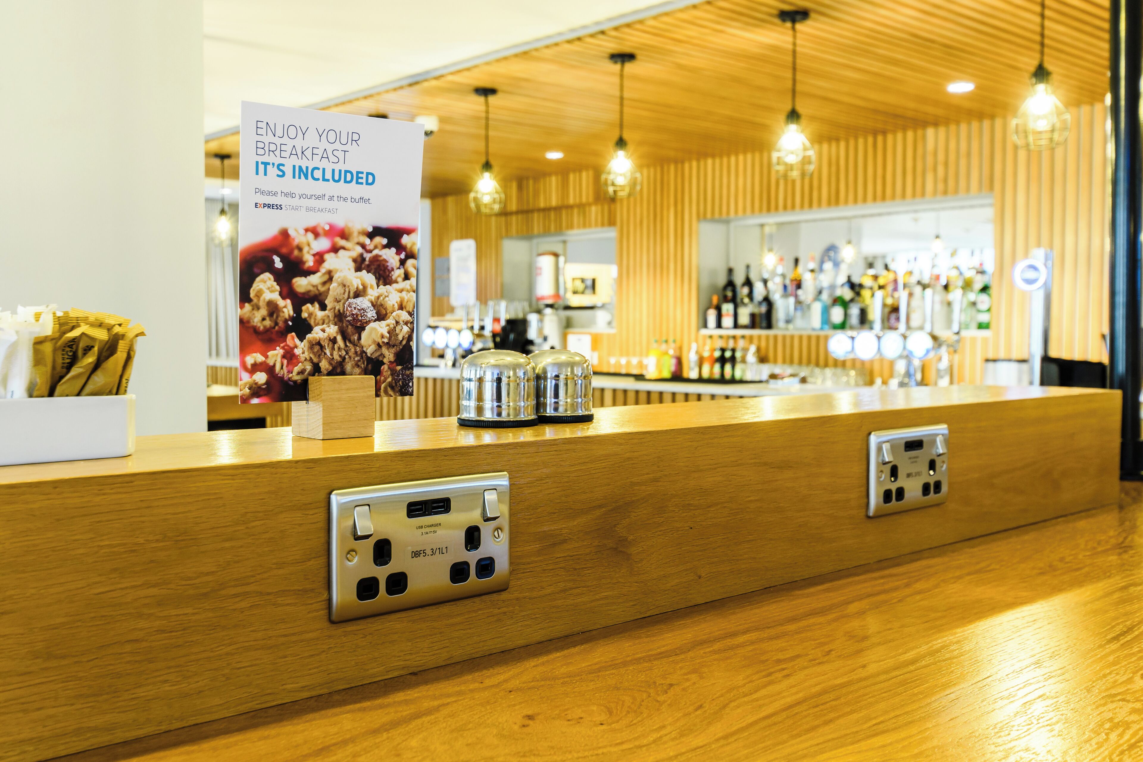 A hotel reception area with a wooden counter, a sign promoting breakfast, and a bar in the background.