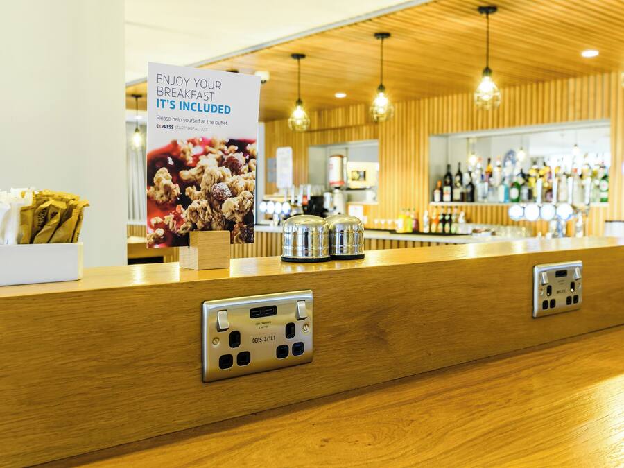 A hotel reception area with a wooden counter, a sign promoting breakfast, and a bar in the background.