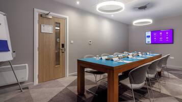 A conference room with a long table, chairs, a whiteboard, and a digital display on the wall.
