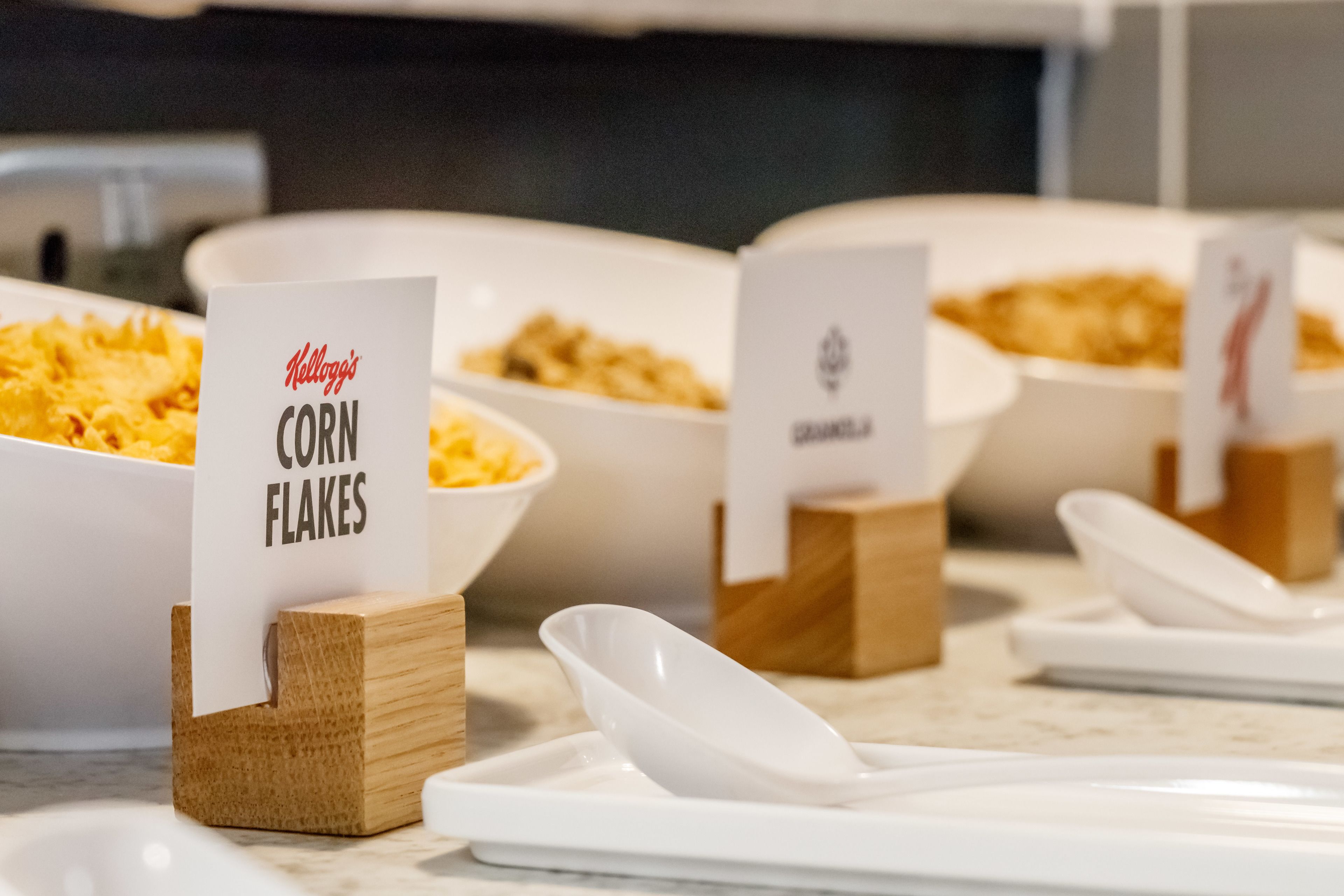Montaje de bufé con tazones de cereal, etiquetados como 'Kellogg's Corn Flakes' y 'Granola'.