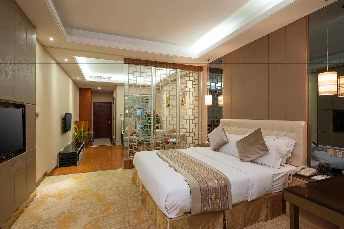 A modern hotel room with a large bed, a desk, a TV, and a bathroom visible through a glass partition.
