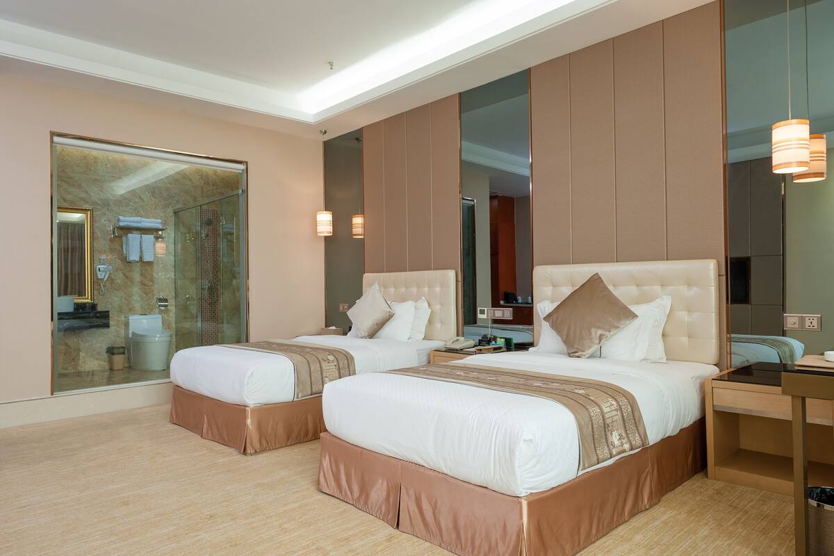 A hotel room with two beds, a glass shower enclosure, a wooden desk, and a large mirror.