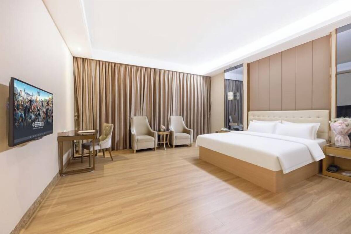 A hotel room with a bed, two armchairs, a small table, a TV, and a large window with curtains.