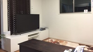 A room with a flat-screen TV on a white stand, a window with a view, and an air conditioning unit on the wall. - Shonan Enoshima Seaside Guest House ANNEX (Kamakura)