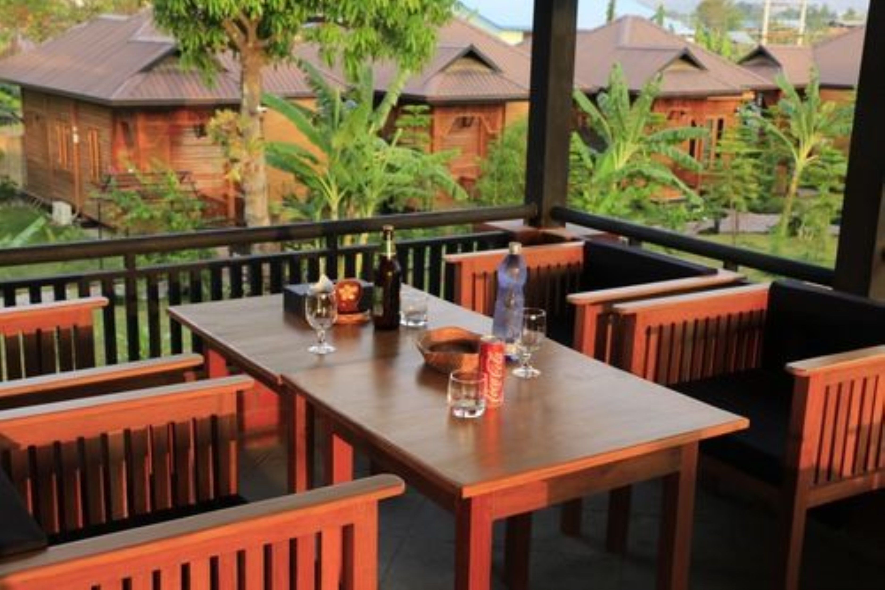 Une table à manger en bois, entourée de chaises, agrémentée de verres et de bouteilles de vin, donnant sur un jardin tropical où se trouvent des bâtiments en bois.