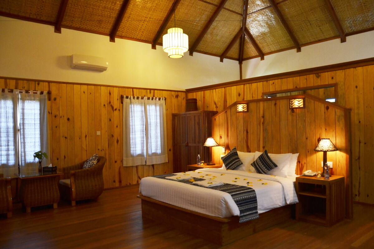 A wooden-themed bedroom with a bed, two bedside tables with lamps, a chair, and a desk.