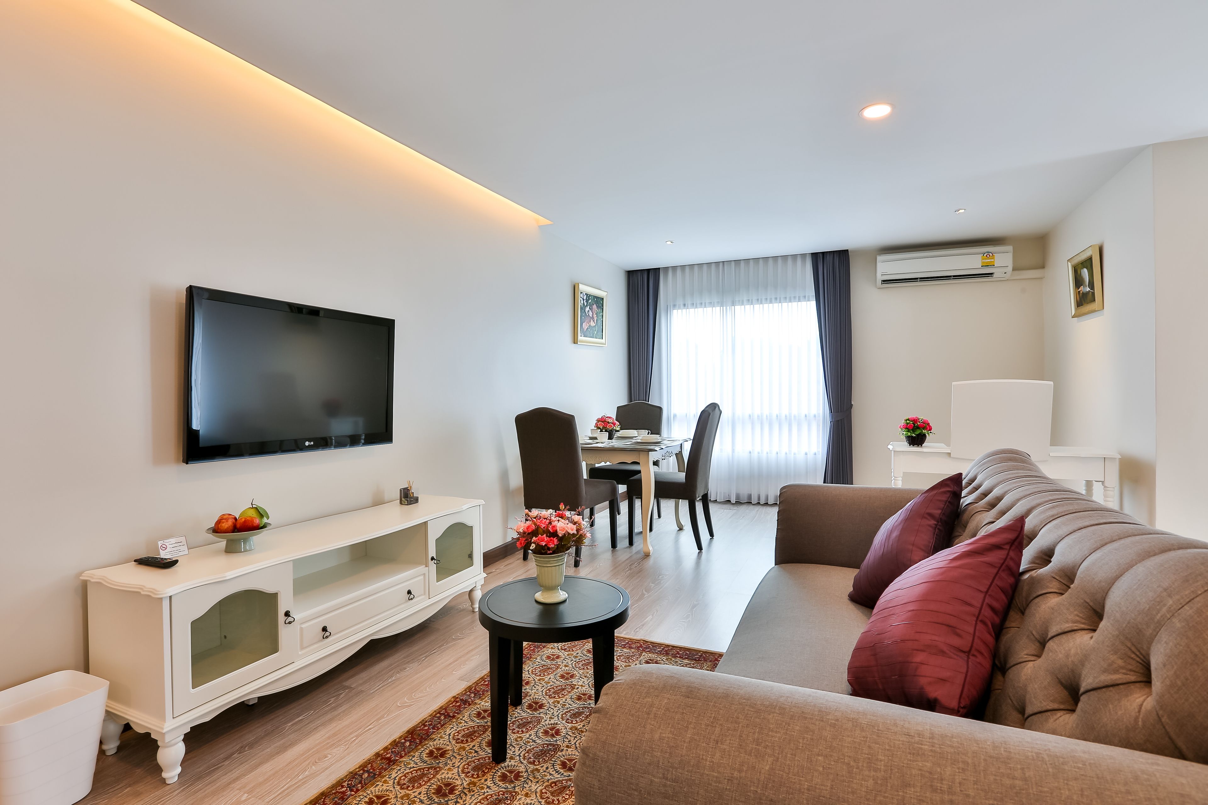 A modern living room with a flat-screen TV, a sofa, a dining area with chairs, and a small side table with flowers.