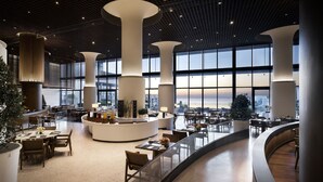 Eine großzügige Lobby mit hohen Decken, großen Fenstern und einer zentralen Sitzecke mit Tischen und Stühlen.