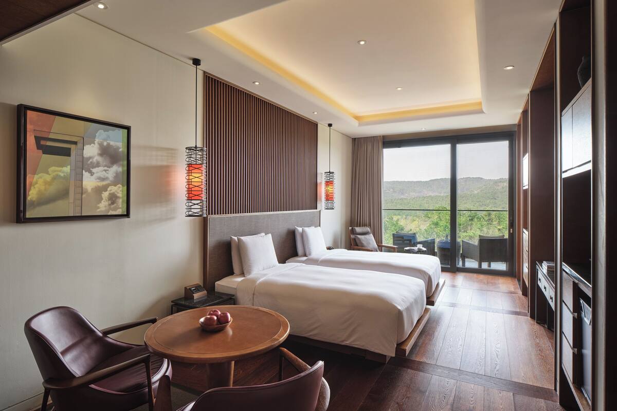 Ein Hotelzimmer mit einem großen Bett, einem Schreibtisch mit Stühlen, einem Esstisch mit Äpfeln und einem Balkon mit Blick auf die Hügel.