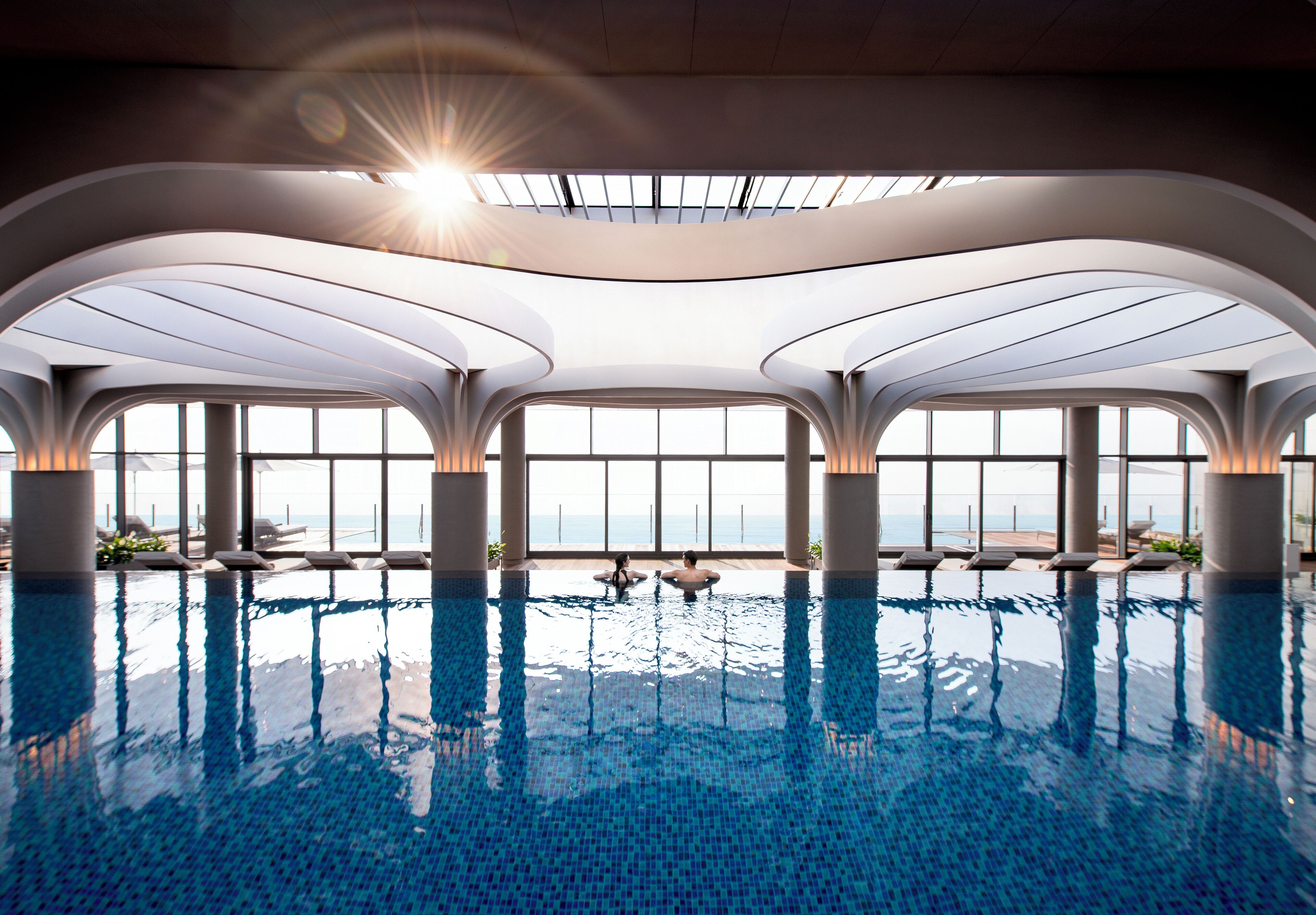 A modern indoor swimming pool with a view of the sea, large windows, and a unique architectural design.